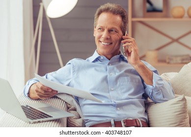 Handsome Middle Aged Businessman Is Using A Laptop, Talking On The Mobile Phone And Smiling While Working At Home