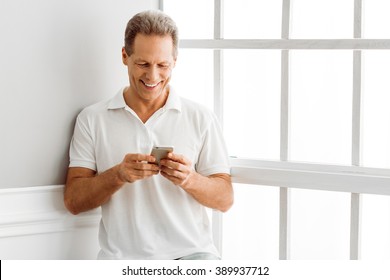 Handsome Middle Aged Man In Casual Clothes Is Using A Smart Phone And Smiling While Sitting Near The Window At Home