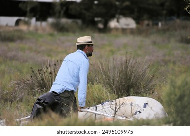 Handsome middle aged man with dark brown skin, black and grey hair and beard, rows a RHIB dinghy through lush plants in wetlands at Burtint UNESCO World Heritage Site. He wears a Panama hat and shirt. - Powered by Shutterstock