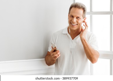 Handsome Middle Aged Man Is Listening To Music Using A Smart Phone And Smiling While Sitting Near The Window At Home