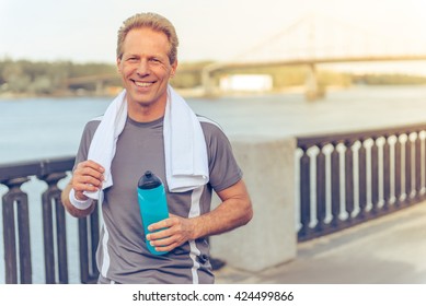 Handsome middle aged man in sports uniform is holding a bottle of water, looking at camera and smiling, resting during morning run - Powered by Shutterstock