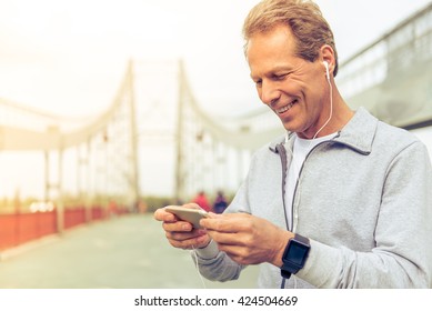 Handsome Middle Aged Man In Sports Uniform And Headphones Is Listening To Music Using A Smartphone And Smiling During Morning Run