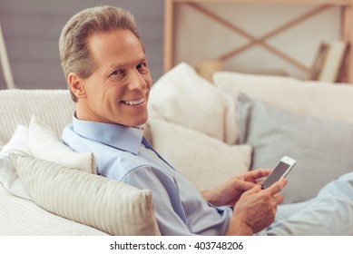 Handsome Middle Aged Man Is Using A Smartphone, Looking At Camera And Smiling While Sitting On Sofa At Home