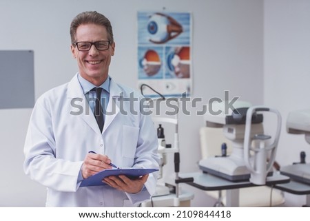 Handsome middle aged ophthalmologist making notes and smiling while standing in his office