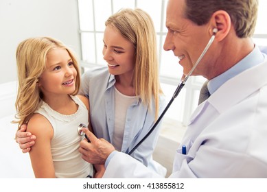 Handsome middle aged pediatrician is sounding little girl lungs. Cute girl and her beautiful mother smiling, sitting in doctors office