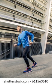 Handsome Middle Aged Serious Man In Blue Black Sports Uniform And Headphones And Fitness Tracker Is Running In The City.