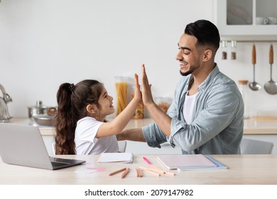 Handsome Middle Eastern Man Dad And Adorable Little Girl Child Sitting At Table, Watching Educational Lesson On Laptop, Taking Notes, Celebrating Success, Giving Each Other High Five, Kitchen Interior