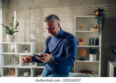 Handsome middle-aged guy using smartphone and a credit card while leaning on a table at home. Gray haired man online shopping or paying bills and rent with an online banking. Finance. Copy space.