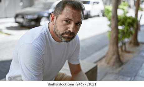 Handsome middle-aged hispanic man with grey hair, wearing a white shirt, sitting outdoors in a blurred urban street setting. - Powered by Shutterstock