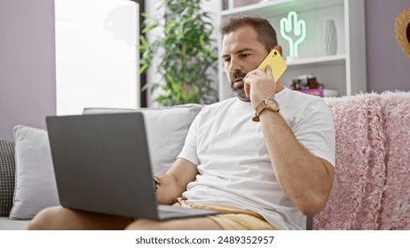 A handsome middle-aged hispanic man with grey hair talking on the phone indoors while relaxing on a sofa with a laptop. - Powered by Shutterstock