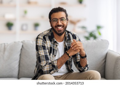 Handsome Middle-eastern Millennial Man Posing At Home, Wearing Casual Outfit And Glasses, Sitting On Couch In Cozy Living Room, Cheerfully Smiling At Camera, Spending Weekend Alone, Copy Space