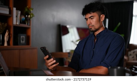 Handsome Millennial Asian Man In His Living Room, Using A Smartphone, Scrolling On Social Media Platform, Chatting With Someone.