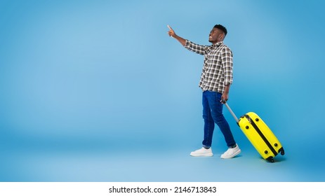 Handsome millennial black guy walking with bright suitcase, pointing at empty space on blue studio background, panorama. Cool male traveler having summer vacation, offering place for tourist agency ad - Powered by Shutterstock