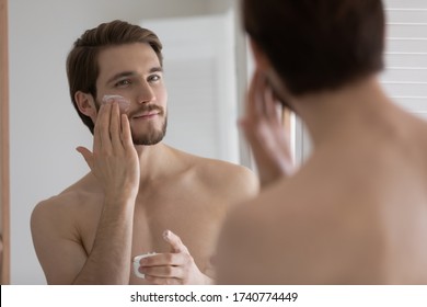 Handsome Millennial Man Look In Bathroom Mirror After Shower Apply Nourishing Facial Cream Or Mask, Young Caucasian Male Use Moisturizing Face Balm For Healthy Glowing Skin, Skincare Concept