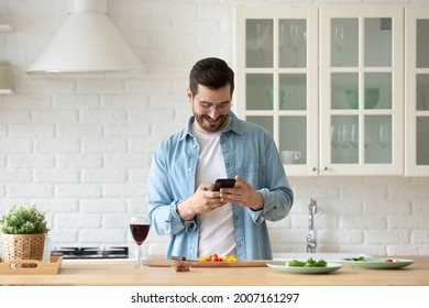 Handsome Millennial Single Man Cooking In Modern Kitchen Holds Smart Phone Searching Recipes On Internet, Looking At Device Screen Smile Make Food Products Delivery Order, Enjoy Easy On-line Services