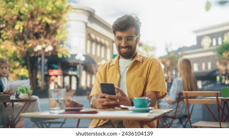 Handsome Multiethnic Man Sitting on a Terrace in a Cafe, Having a Cup of Coffee with Pastry. Happy Indian Man Connecting with Friends Online, Replying to Social Media Posts and Emails on a Smartphone - Powered by Shutterstock