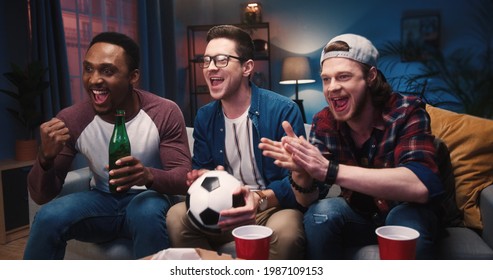 Handsome multi-ethnic young joyful males sitting on couch at home in evening drinking from paper cups watching football event on TV cheering for favorite team, leisure, entertainment, fans concept - Powered by Shutterstock