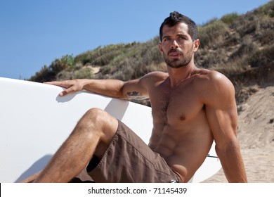 Handsome muscular dark haired surfer sitting on the sand next to his board, wearing a brown bathing suit.
