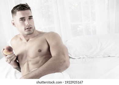 Handsome muscular man lying shirtless in bed revealing pecs and defined sixpack abs, holding a peach and looking at camera