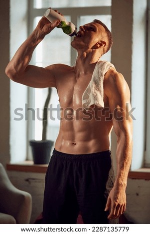 Handsome muscular man with relief strong body, standing shirtless, drinking water after training session at home on daytime. Concept of sportive lifestyle, body and health care, fitness, health