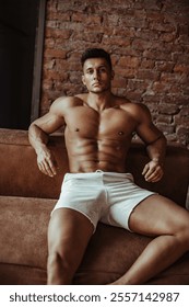 Handsome muscular man in white sports shorts posing in loft interior. Artistic portrait of male fitness model in shorts sitting on sofa. Naked brunette with six pack abs posing in studio.