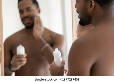 Handsome muscular mixed race Black young man applying aftershave lotion, balm on soft stubble, stylish beard at mirror, holding plastic bottle, touching face. Male beauty care procedures concept