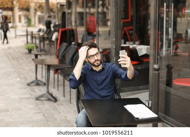 Handsome Muslim Male Person Making Selfie By Smartphone At Street Cafe Table. Attractive Man Dressed In Blue Shirt Using Mobile Phone As Mirror. Concept Of Taking Photos With Front Camera.