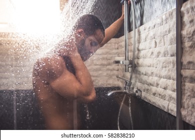 Handsome naked Afro American man is leaning on the wall while taking shower in bathroom