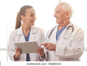 Handsome Old Doctor And Beautiful Young Female Doctor In White Medical Coats Are Using A Digital Tablet, Talking And Smiling