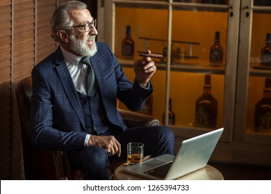 Handsome Old Male Pensioner Dressed In Smart Formal Style Using Laptop And Smoking While Sitting On Couch At Luxury Home Interior With Cabinet With Expensive Alcohol.