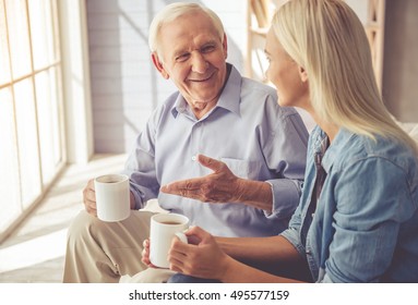 Handsome old man and beautiful young girl are  drinking tea, talking and smiling while sitting on couch at home