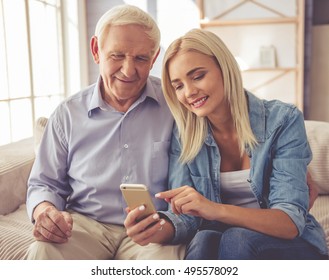 Handsome old man and beautiful young girl are using a smartphone, talking and smiling while sitting on couch at home