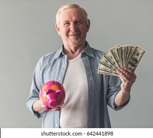 Handsome Old Man In Casual Wear Is Holding A Piggy Bank And Money, Looking At Camera And Smiling, On Gray Background