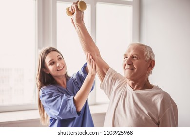 Handsome old man is doing exercises with dumbbells and smiling, in hospital ward. Attractive nurse is helping him