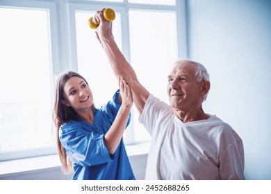 Handsome old man is doing exercises with dumbbells and smiling, in hospital ward. Attractive nurse is helping him - Powered by Shutterstock