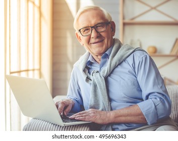 Handsome Old Man Dressed In Smart Casual Style And Eyeglasses Is Using A Laptop, Looking At Camera And Smiling While Sitting On Couch At Home