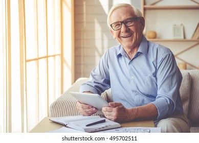 Handsome Old Man In Eyeglasses Is Using A Digital Tablet, Looking At Camera And Smiling While Working At Home