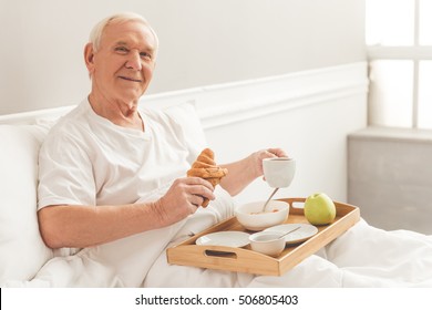 Handsome Old Man Is Holding A Tray With Breakfast, Looking At Camera And Smiling While Lying In Bed