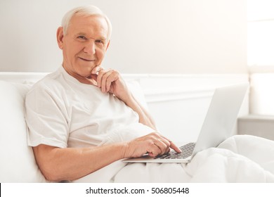 Handsome Old Man Is Using A Laptop, Looking At Camera And Smiling While Lying In Bed