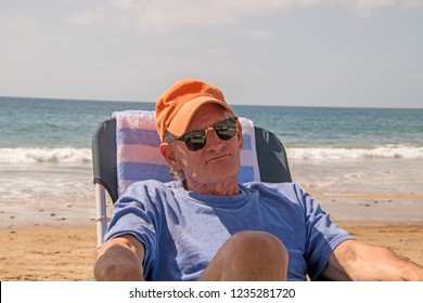 Handsome Older Baby Boomer Man Wearing An Orange Cap Sitting On Beach Chair By The Ocean With A Frown On His Face