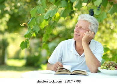 Handsome Older Man Sitting At A Table At Home On The Veranda With Book