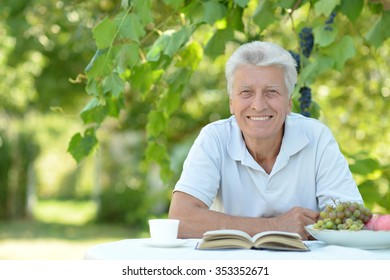 Handsome Older Man Sitting At A Table At Home On The Veranda With Book