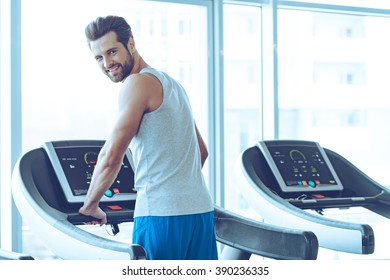 Handsome On Treadmill. Rear View Of Young Handsome Man In Sportswear Standing On Treadmill In Front Of Window At Gym And Looking At Camera With Smile