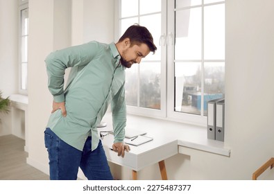 Handsome oung man suffering from backache in ofiice. Office worker standing leaning table touching his painful lower back, man feeling pain or spinal spasm. Sedentary life, office syndrome concept