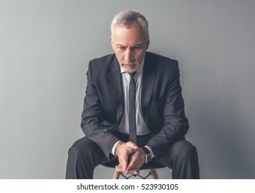 Handsome pensive mature businessman in formal suit is looking down while sitting on chair, on gray background