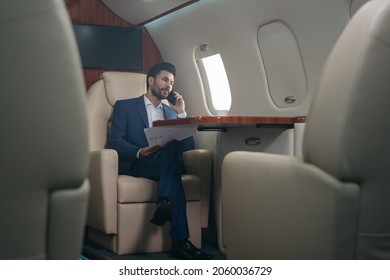 Handsome pensive middle eastern businessman talking on mobile phone, holding financial report working with documents sitting in plane. Confident entrepreneur flying private jet, successful business - Shutterstock ID 2060036729