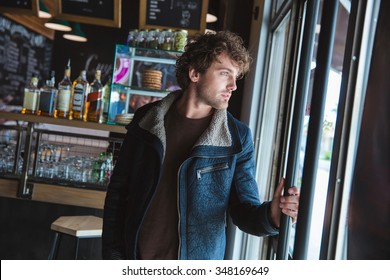 Handsome Pensive Young Curly Man In Black Jacket Looking Through Glass Door In Cafe