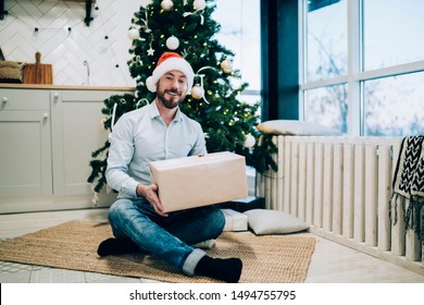 Handsome Pleased Man Sitting Near Christmas Tree And Unwrapping Carton Box With Gift In Decorated Room At Christmas Looking At Camera