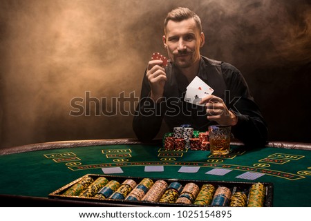 Handsome poker player with two aces in his hands and chips sitting at poker table in a dark room full of cigarette smoke.