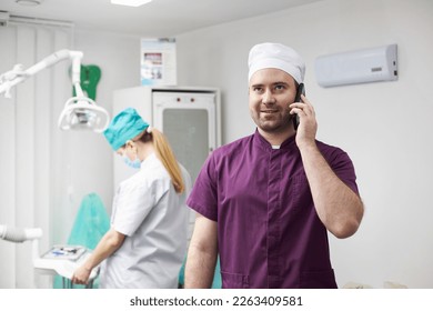 Handsome positive bearded Caucasian young man dentist doctor, smiling while talking on mobile phone in a dentistry clinic. Dentist assistant on the background. People. Service. Consumerism. Healthcare - Powered by Shutterstock